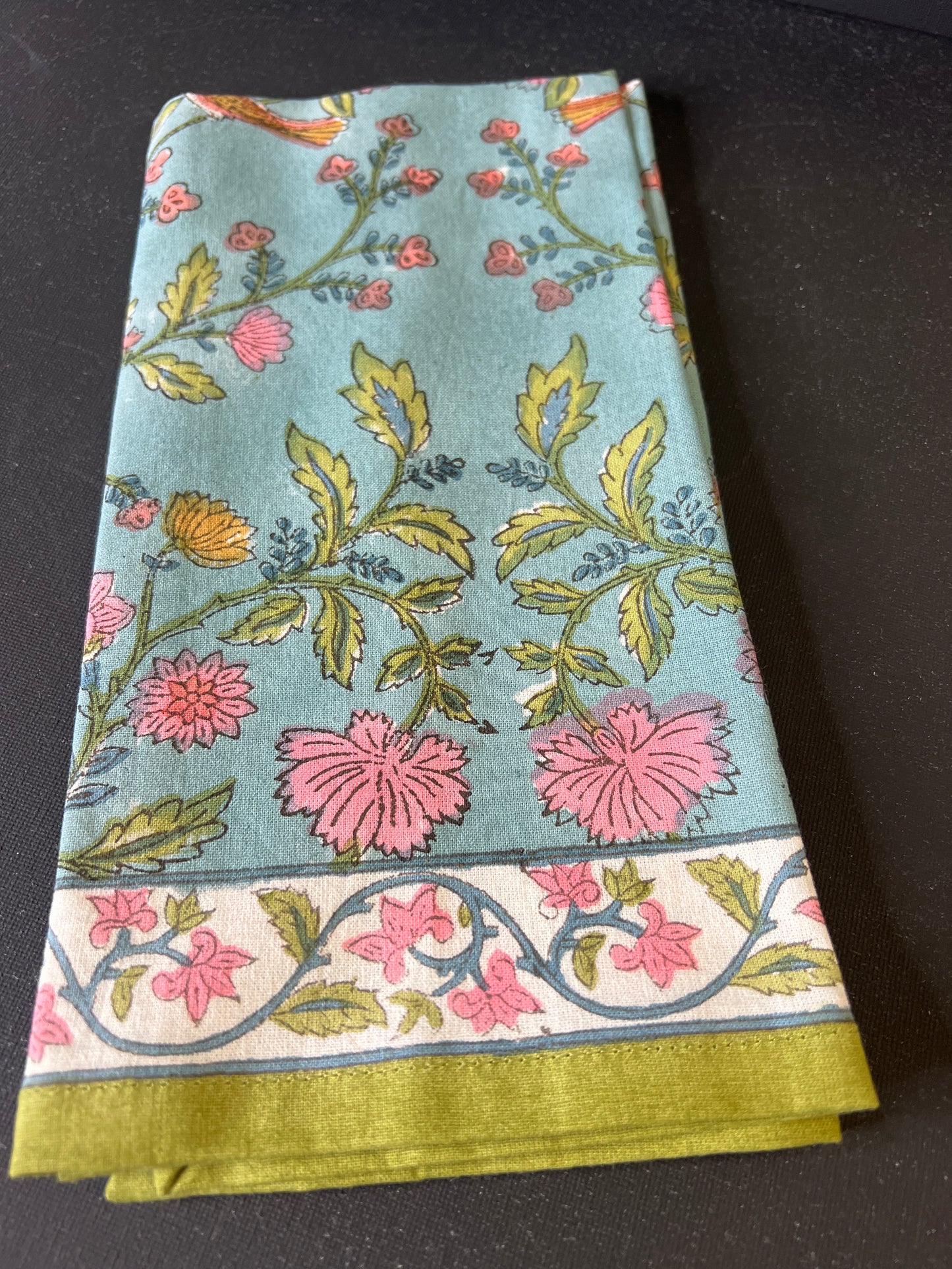 Floral patterned fabric with pink flowers and green leaves on a light blue background, folded and placed on a dark surface.