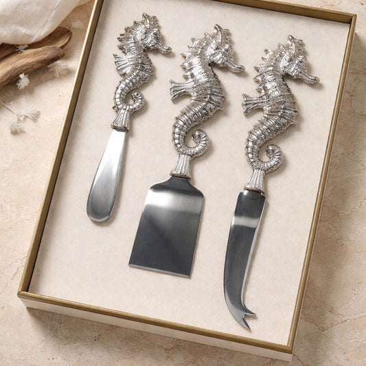 Set of three seahorse-shaped cheese knives in a box on a beige surface.