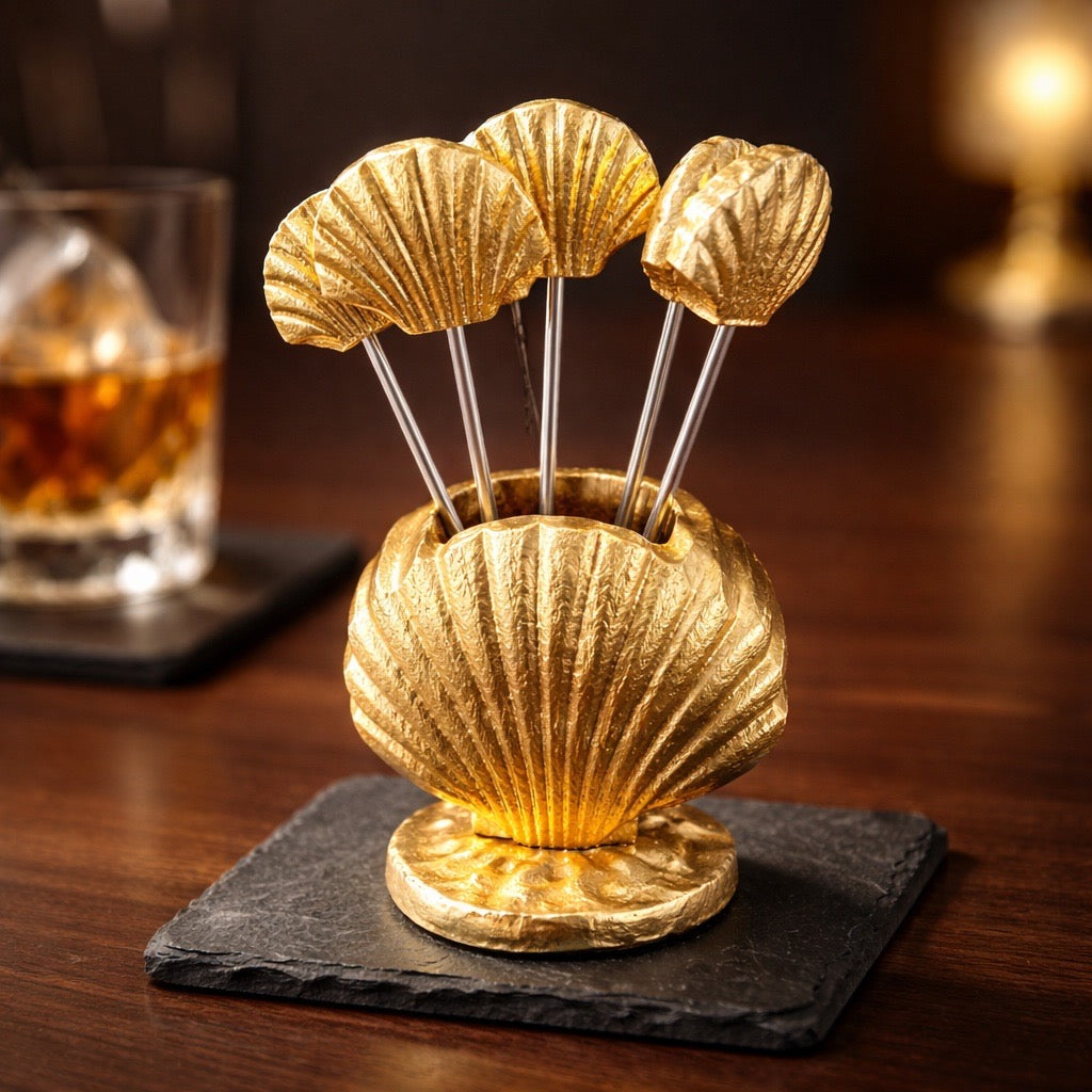 Gold shell-shaped cocktail stirrer set on a dark coaster with a blurred drink in the background.